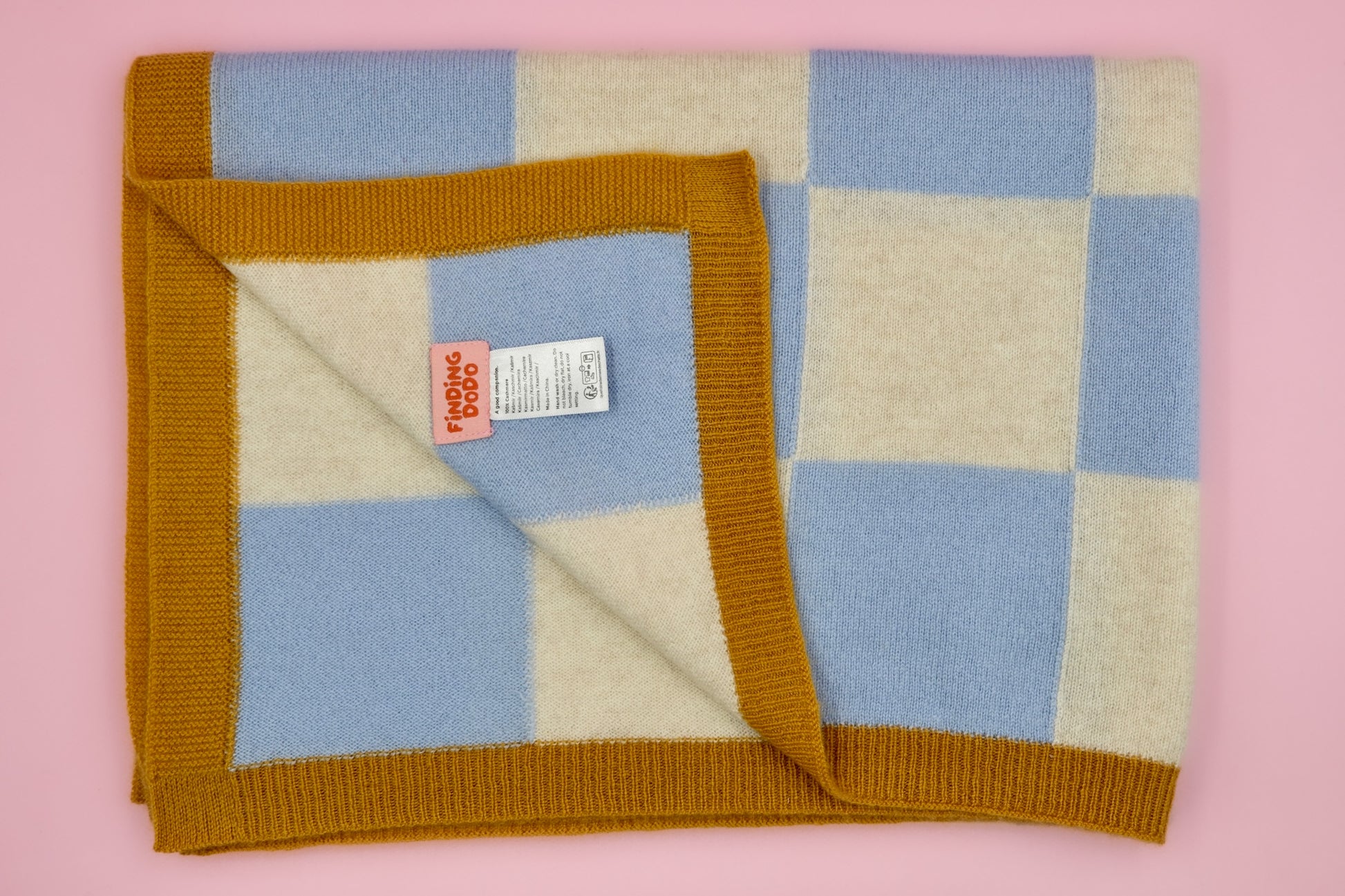 Rechteckig gefaltete Finding Dodo Kaschmirdecke Ari in Beige mit braunem Rand und sichtbarem Markenlabel vor rosa Hintergrund.