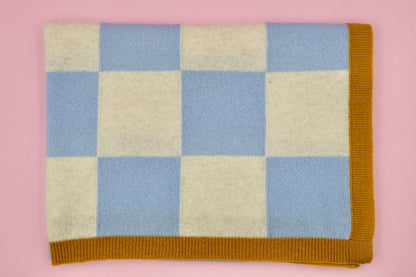 Beige-blau karierte Finding Dodo Kaschmirdecke mit braunem Rand, rechteckig gefaltet vor einer rosa Wand.