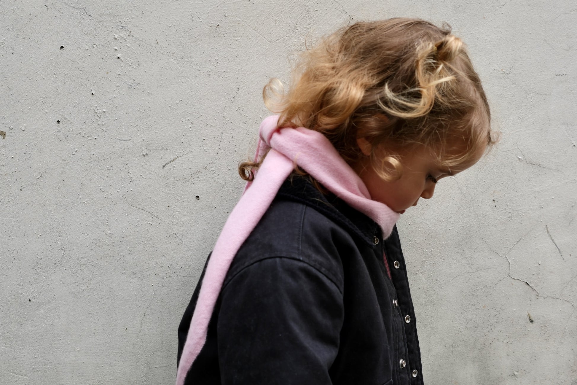 Kleines Mädchen mit Locken in blauer Jeansjacke trägt das rosa Finding Dodo Kaschmir-Dreiecktuch, Seitenansicht vor grauem Hintergrund.